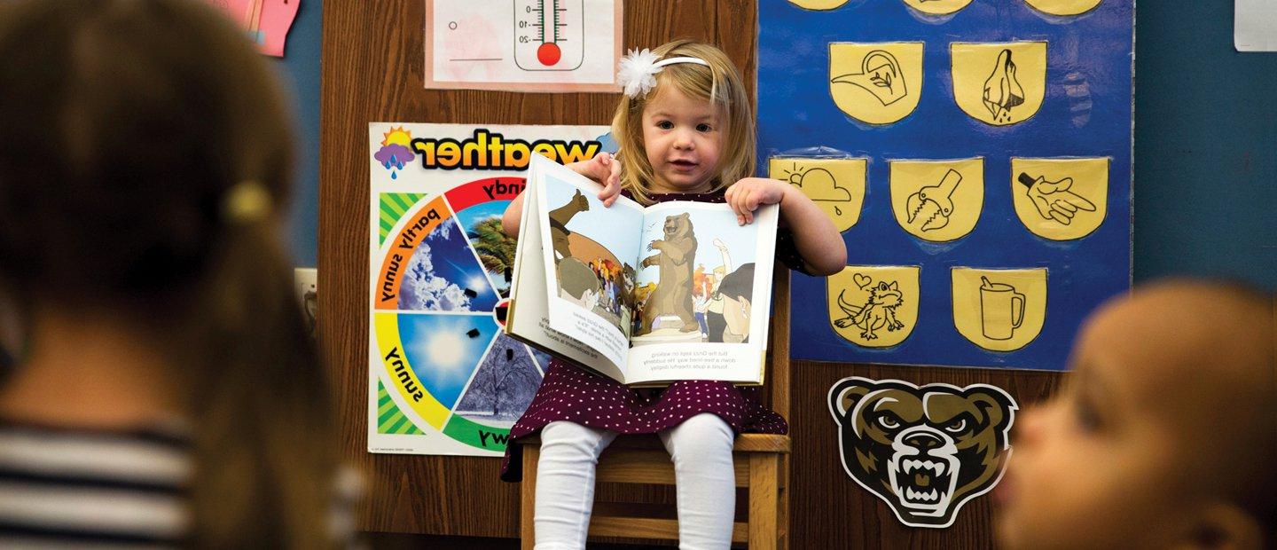 Young girl seated at the front of a classroom, showing her classmates a book.