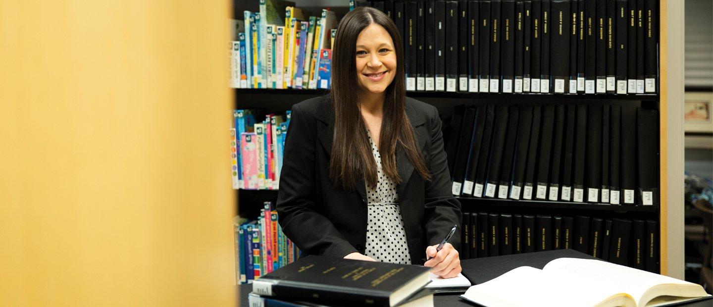 A woman standing at a table with open books, with shelves of books behind her.